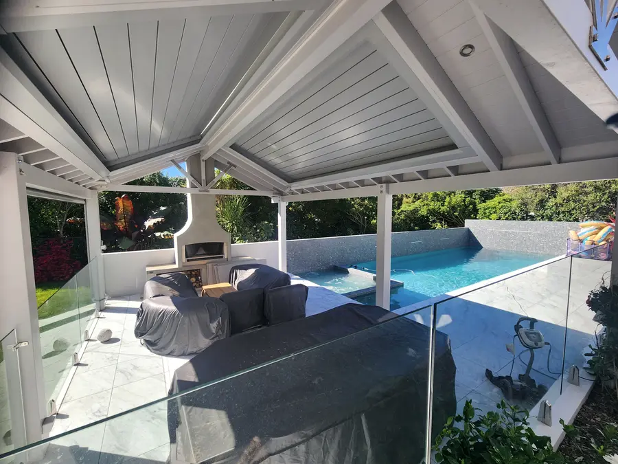A white pergola with a timber-lined ceiling and outdoor fireplace, looking out over a tiled pool